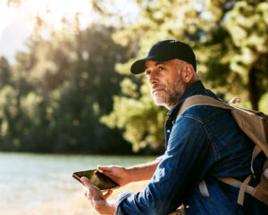 A senior man with a backpack sits by a lakeside holding a tablet, symbolizing outdoor activity and continued independence after a glaucoma diagnosis